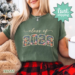 May include: Green t-shirt with the text "class of 2026" in a script font. The numbers "2026" are filled with a floral pattern. The shirt is a moss green color. A "fast shipping" graphic is in the upper right corner.