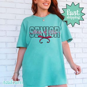 May include: Seafoam green t-shirt with the text "SENIOR Class of 2024" in black and pink, and a pink bow graphic. The shirt has short sleeves and a relaxed fit. A green starburst graphic with the words "fast shipping" is in the upper right corner.