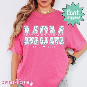 Könnte beinhalten: Ein leuchtend pinkfarbenes T-Shirt mit dem Schriftzug "MAMA MUM" in weißen Blockbuchstaben, die mit einem blauen Blumenmuster gefüllt sind. Der Text "CUSTOM UNIVERSITY" steht über den Buchstaben, und "EST. 2026" darunter, mit einem Herzsymbol. Eine grüne Stern-Grafik sagt "fast shipping".