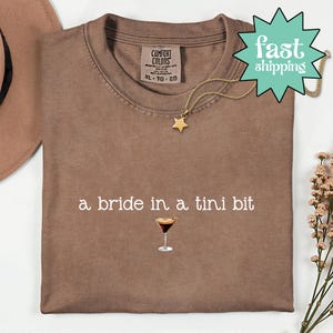 May include: A brown Comfort Colors t-shirt with the text "a bride in a tini bit" and a cocktail graphic. A gold star necklace and a "fast shipping" tag are also visible. The shirt is XL size.