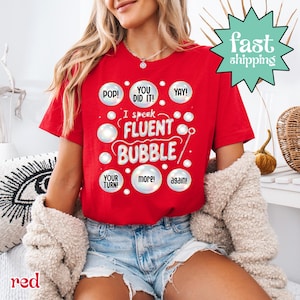 May include: A red t-shirt with the text "I speak FLUENT BUBBLE" in white, surrounded by bubble graphics and phrases like "POP!" and "YAY!". The shirt also has the words "YOUR TURN!", "more!", and "again!" in white bubbles. A "fast shipping" graphic is in the top right.