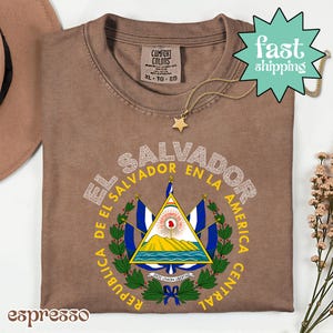 a brown t-shirt with a graphic design of the coat of arms of El Salvador, along with a star and a necklace.