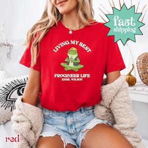 以下が含まれることがあります： 「LIVING MY BEST」の文字と、ヘルメットをかぶりコーヒーカップを持ったカエルのイラストが描かれた赤いTシャツ。「FROGINEER LIFE ENGR. WILSON」の文字が下にあります。緑色のスターバーストグラフィックには「fast shipping」と書かれています。