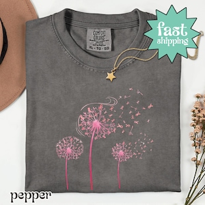 以下が含まれることがあります： ピンクのタンポポのデザインとピンクのリボンが付いた、濃いグレーのTシャツ。Tシャツはクルーネックと半袖です。金の星のペンダントネックレスがTシャツの上にかけられています。「pepper」の文字が下部に印刷されています。