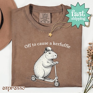Könnte beinhalten: Espressofarbenes T-Shirt mit dem Text "Off to cause a kerfuffle" und einem Capybara auf einem Roller. Das Shirt hat ein Comfort Colors-Etikett und eine goldene Sternkette. Ein "fast shipping"-Tag ist sichtbar.