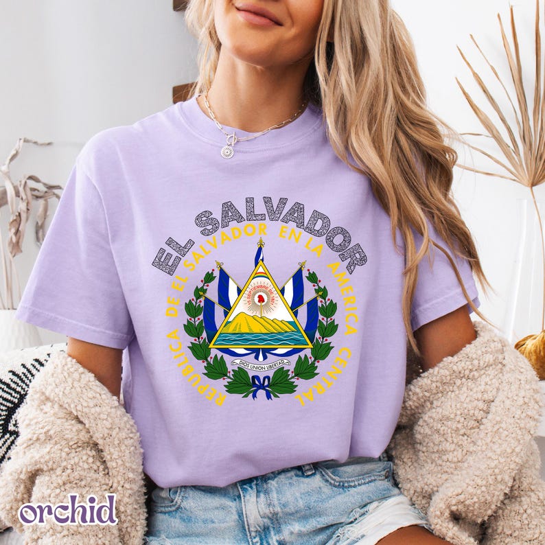 A woman wearing a purple t-shirt with the text "El Salvador" and a graphic of the Salvadoran flag, along with the text "El Salvador de la América" and "RePUBLICA DE EL SALVADOR EN LA AMERICA".