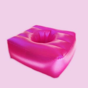 Op de afbeelding: Een felroze opblaasbaar kussen met een centrale cirkelvormige opening. Het kussen is vierkant van vorm en heeft een geribbelde oppervlakte voor comfort. Dit opvallende item is ontworpen voor ondersteuning.