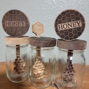Puede incluir: Tres frascos de vidrio con tapas de madera y cucharas para miel. Las tapas están grabadas con un patrón de panal y la palabra "Honey".