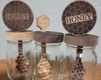 Puede incluir: Tres frascos de vidrio con tapas de madera y cucharas para miel. Las tapas están grabadas con un patrón de panal y la palabra "Honey".