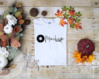 May include: White t-shirt with a black camera lens and the text "Mockup Glam" printed on the front. The t-shirt is laying flat on a wooden surface with fall-themed decorations.