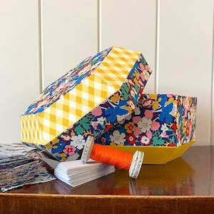 Pode incluir: Uma caixa de tecido floral com uma tampa de vichy amarela e branca. A caixa está aberta e há um carretel de fio laranja e uma pilha de cartões brancos sobre uma mesa de madeira.