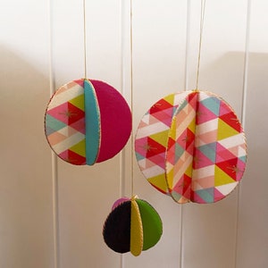 Puede incluir: Tres adornos de papel coloridos que cuelgan del techo. Los adornos están hechos de círculos de papel con patrones geométricos en rosa, amarillo, azul y verde. Los adornos están suspendidos de una cuerda dorada.