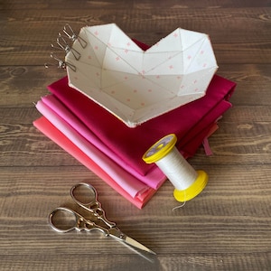 May include: A white heart-shaped fabric bowl with a geometric pattern, a spool of white thread with a yellow bobbin, a pair of gold scissors, and a stack of pink fabric.