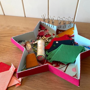 Op de afbeelding: Een roze stervormige stoffen doos met een witte en bloemige voering. De doos bevat naai benodigdheden, waaronder een spoel wit garen, een klein glazen flesje, een schaar en verschillende stukken stof in rood, groen en blauw.