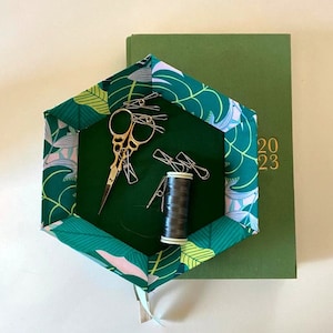 May include: A hexagonal fabric tray with a green and pink tropical leaf pattern. The tray contains a pair of gold scissors, a spool of black thread, and several gold paper clips. The tray is sitting on top of a green journal with the numbers 2023 printed in gold on the cover.