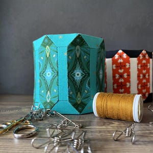 May include: A teal fabric box with a geometric pattern, a spool of gold thread, and silver binder clips on a wooden surface.