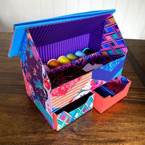 Puede incluir: Un organizador de almacenamiento en forma de casa cubierto con papel estampado colorido con diseños florales, geométricos y de rayas. Cuenta con un techo azul brillante, un compartimento superior abierto con carretes de hilo y dos cajones extraíbles.