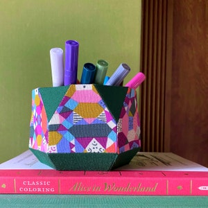 Könnte beinhalten: Ein farbenfroher geometrischer Stoff-Bleistifthalter mit grüner Basis, gefüllt mit violetten, rosa und grünen Markern. Der Halter steht auf einem Buch mit dem Titel "Alice im Wunderland: Klassisches Ausmalen".