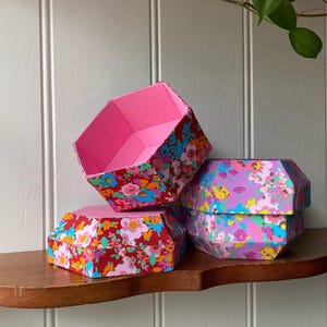 May include: Three hexagonal decorative boxes with floral patterns. The boxes are in shades of red, pink, and purple, with colorful flower designs. The boxes are stacked on a wooden shelf against a white wall.