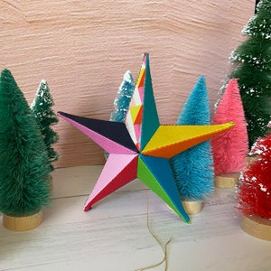 Puede incluir: Un adorno de estrella colorido y de múltiples puntas hecho de tela con una variedad de patrones y colores, incluyendo rosa, amarillo, azul, verde y negro. La estrella está colgada de una cuerda y está rodeada de pequeños árboles de Navidad artificiales.