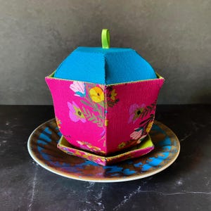 Puede incluir: Un juego de tetera y platillo de tela rosa y verde con un patrón floral. La tetera tiene una tapa de tela azul con una cinta verde.