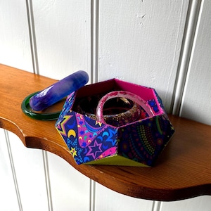 Może przedstawiać: Geometryczne pudełko z tkaniny z wzorem paisley i gwiazd, zawierające kilka bransoletek. Fioletowa i zielona bransoletka leżą obok pudełka na drewnianej półce. Pudełko ma różowe wnętrze i zieloną podstawę.