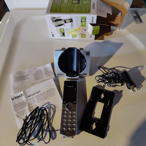 May include: A silver and black VTech cordless phone system, including a base station, handset, charging cradle, and paperwork. The phone has a numeric keypad and a black screen. The box is partially open.