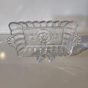 May include: Clear glass rectangular serving dish with a decorative floral and dot pattern. The dish has a scalloped edge and small handles on each side. The dish is supported by a clear plastic stand.