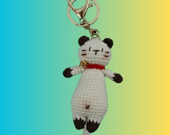 May include: A handmade white crochet cat keychain with brown accents on the ears, paws, and tail. The cat has a red collar with a small gold bell and a gold-colored keychain ring. The background is a gradient of blue, green, and yellow.