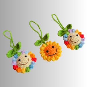 May include: Three crocheted sunflower ornaments with smiling faces. The sunflowers have yellow, orange, and rainbow-colored petals, green leaves, and green hanging loops. The ornaments are handmade and have a cheerful, whimsical design.