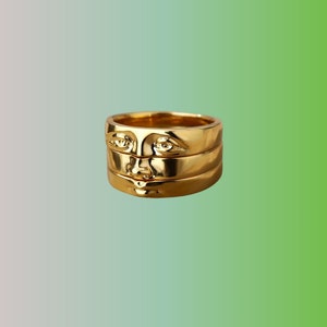 May include: A gold ring with a layered design featuring a face with eyes, nose, and mouth.