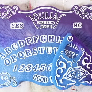 Puede incluir: Una tabla de Ouija morada y azul con letras blancas y una planchette blanca con un diseño de ojo azul. La tabla dice "Ouija Mystifying Oracle" y tiene las letras A-Z, los números 1-9, 0, y las palabras "Sí", "No", y "Adiós".
