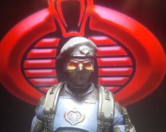 May include: A blue and black action figure of a soldier wearing a blue and gray helmet and a black mask. The figure is standing in front of a red Cobra symbol.