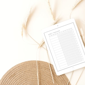 May include: A white digital tablet with a black border displaying a habit tracker. The tracker has a title "Habit Tracker" and columns for each day of the week. The tablet is on a white surface with dried wheat stalks and a woven straw placemat.