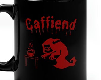 May include: Black ceramic coffee mug with a red graphic of a spooky figure and the word "Caffiend" in a dripping font. The design includes a cauldron and a table. The mug has a curved handle.
