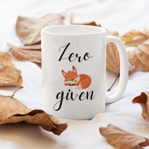 May include: White ceramic mug with a black script that reads "Zero given" and a cartoon fox illustration. The fox is orange and white, curled up asleep. The mug has a white handle.
