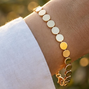 May include: A gold-colored bracelet featuring a series of small, circular discs linked together. The bracelet is worn on a wrist, with a white shirt sleeve visible in the frame. The background is blurred, with warm, natural tones.