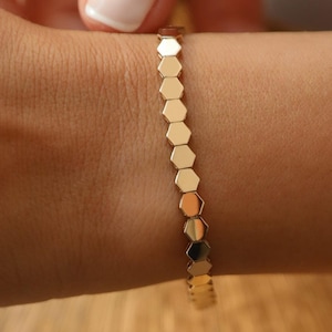 May include: A gold bracelet featuring a series of connected hexagonal links. The bracelet is shown on a wrist, with a hand gently touching it. The bracelet's design is simple and elegant, reflecting a modern aesthetic.