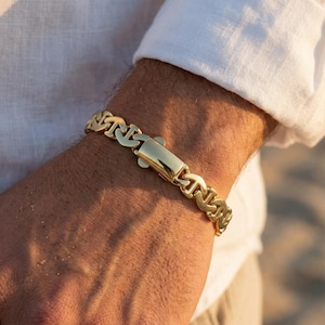 May include: A polished gold-tone chain bracelet featuring interlocking anchor-shaped links and a smooth, rectangular box clasp closure. The bracelet has a high-shine metallic finish and is worn around the wrist, showcasing the detailed link design.