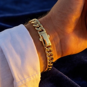 May include: A gold curb chain bracelet with a secure clasp. The bracelet is worn on a wrist, contrasting with a white shirt sleeve and a dark blue background. The links are thick and shiny.