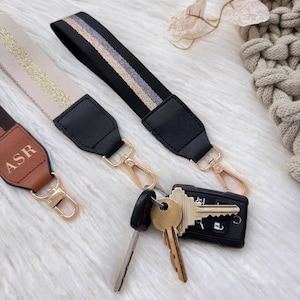 May include: Close-up of three purse straps with gold-colored hardware. One strap is black with gold and silver stripes, another is beige with gold stripes, and the third is brown with the initials "ASR". Keys are attached to one strap.