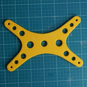 Puede incluir: Un componente amarillo brillante en forma de estrella con múltiples agujeros circulares y bordes redondeados. El objeto se encuentra sobre un fondo de cuadrícula verde azulado, lo que sugiere un contexto de artesanía o ingeniería.