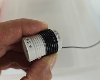 May include: A white plastic coil with a black rubber band wrapped around it. The coil has multiple holes and is connected to a black wire.