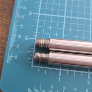 Puede incluir: Dos varillas roscadas plateadas, probablemente para construcción o reparación. Las varillas están paralelas sobre una esterilla de corte azul con líneas de cuadrícula y medidas. Las varillas tienen un acabado liso y brillante. Medidas en centímetros.