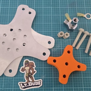 以下が含まれることがあります： 複数の穴がある銀色の金属製の星型プレート、オレンジ色の星型パーツ、ボルト、ナット、そして「LT.DUDE」の文字と漫画のキャラクターが描かれたステッカーなど、さまざまなハードウェアコンポーネントの詰め合わせ。
