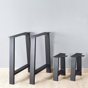 May include: A set of four matte black metal table legs featuring a trapezoidal frame design. The set includes two tall, wide legs and two shorter, narrower legs, each constructed with a flat top mounting plate and a sturdy rectangular base bar.