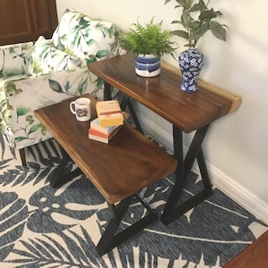 May include: A wooden bench and a wooden side table with black metal legs. The bench and table are both made of natural wood with a live edge. The table has a vase with blue and white flowers and a potted plant on top. The bench has a mug and books on top.