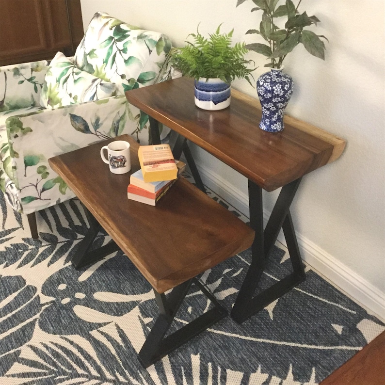 May include: A wooden bench and a wooden side table with black metal legs. The bench and table are both made of natural wood with a live edge. The table has a vase with blue and white flowers and a potted plant on top. The bench has a mug and books on top.