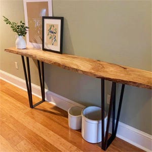 May include: A long console table featuring a natural live-edge light wood slab top supported by two black metal U-shaped legs. The table surface holds a white ceramic vase with greenery and two framed prints, while two white ceramic crocks sit below.