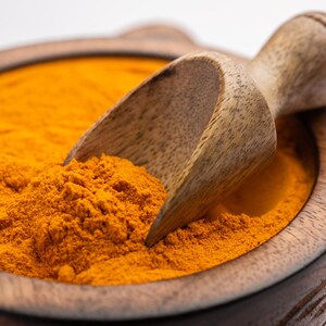 K&ouml;nnte beinhalten: Eine Holzschale gef&uuml;llt mit leuchtend orangefarbenem Kurkuma-Pulver. Eine Holzschaufel ist teilweise in das Pulver eingetaucht.